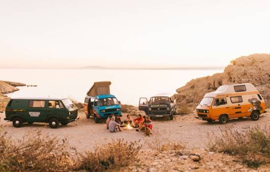 Plusieurs vans aménagés stationnés près du littoral autour d’un feu de camp dans un paysage naturel.