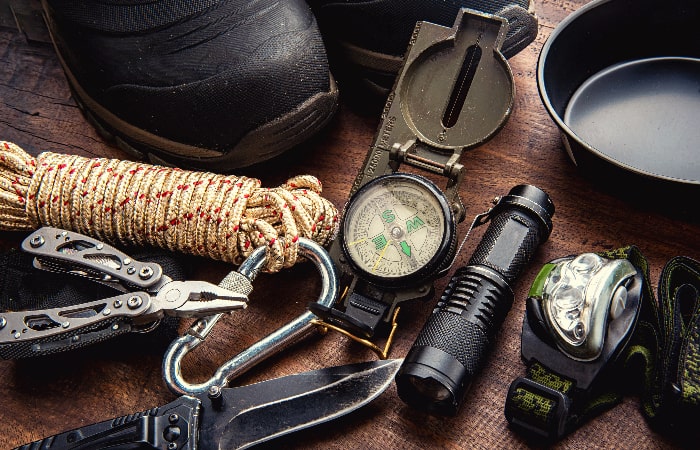 Équipements de randonnée posés sur une table en bois, incluant boussole, corde, couteau multifonction, lampe frontale et chaussures.