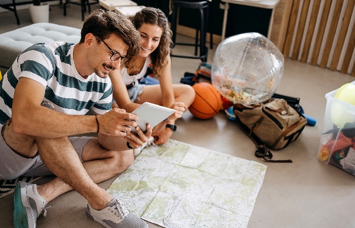 Jeune couple assis au sol en train de planifier un itinéraire avec une carte et une tablette, entouré d’affaires de voyage.