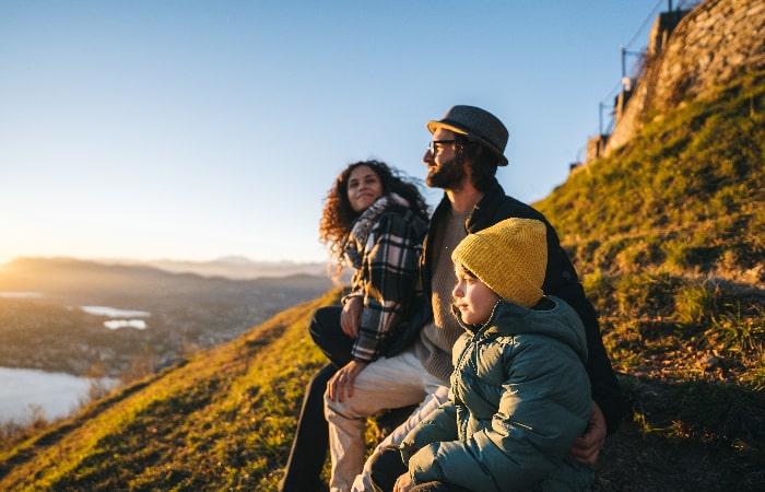 Famille assise sur une colline au coucher du soleil, admirant le paysage naturel en contrebas.